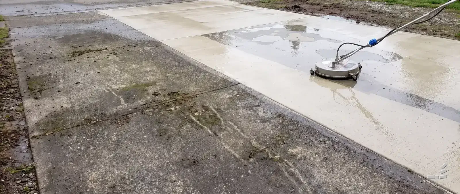 Photograph-style driveway apron, algae-stained concrete on the near side of one diagonal and bare wet clean concrete beyond it, a surface cleaner resting on the clean half